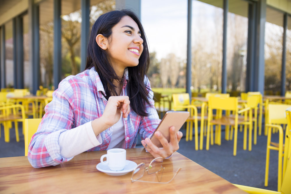 young lady sitting outside cafe smiling and holding her mobile phone